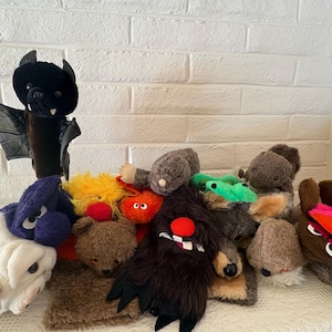 May include: A collection of various plush hand puppets including a black bat with wings, a white bulldog, a brown bear, a shaggy dark brown monster with a red nose, a bright orange creature, a yellow fuzzy puppet, and several other animal figures.