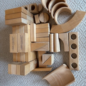 May include: A collection of natural light-colored wooden building blocks in various geometric shapes, including arches, rectangles, triangles, and a long piece with four circular cutouts. Several blocks are stamped with the text CHILDCRAFT MADE IN U.S.A.