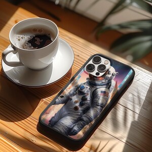 Könnte beinhalten: Schwarze Smartphone-Hülle mit einem Astronauten-Design vor einem Weltraum- und Wolkenhintergrund. Eine weiße Kaffeetasse und Untertasse stehen auf einer Holzoberfläche. Im Hintergrund ist eine Pflanze zu sehen.