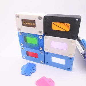 May include: A collection of rectangular electronic display housings in white, black, and blue, featuring colored acrylic windows in orange, green, red, and clear. Two small, separate rectangular covers in blue and pink are placed in the foreground.
