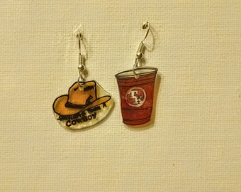 May include: A pair of earrings featuring a red solo cup with a white "TX" logo and a cowboy hat with the text "Shouldn't've Been a Cowboy".