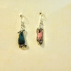 May include: A pair of dangle earrings featuring cartoon characters with the word "Yip!" printed on them. One earring is blue and the other is pink.