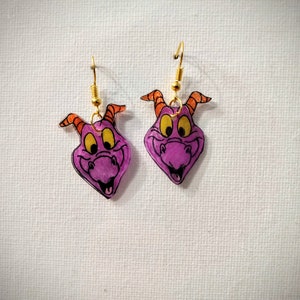 May include: A pair of purple dragon earrings with orange horns and gold hooks. The dragon's face is drawn with black lines and has yellow eyes.