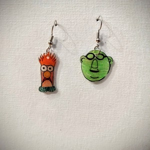 May include: A pair of earrings featuring cartoon characters. One earring is green and depicts a character with glasses. The other earring is orange and depicts a character with red hair.