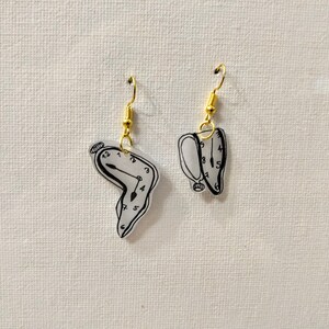 May include: A pair of gold earrings with a white, melted clock face design. The clock face has black numbers and hands.
