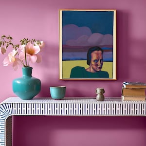May include: A teal blue ceramic vase with pink flowers sits on a white and black patterned console table. A framed painting of a person with a dark blue background hangs on a pink wall. A small ceramic bowl and a ceramic animal figurine are also on the table.