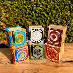 Puede incluir: Una colección de cajas de libros decorativas con temática de fantasía. Las cajas presentan diseños intrincados con detalles dorados, azules, rojos y verdes. Cada caja tiene un emblema único y un cierre de cinta.