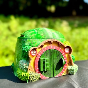 May include: A green and yellow hobbit house with a red and green door. The house is made of ceramic and has a detailed design.