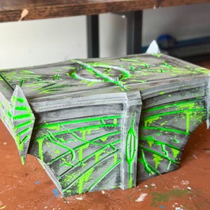 May include: A gray and black decorative box with neon green paint splatters and accents. The box has a geometric design with pointed edges and a circular design on top. The box appears to be 3D printed.