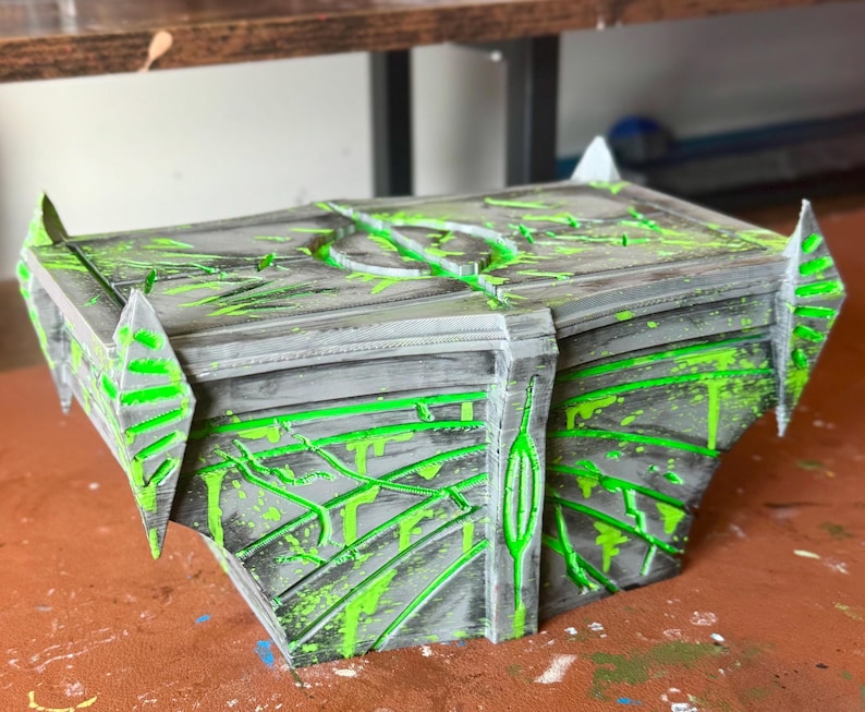 May include: A gray and black decorative box with neon green paint splatters and accents. The box has a geometric design with pointed edges and a circular design on top. The box appears to be 3D printed.