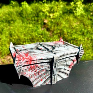 May include: A gray and white decorative box with red splatter paint details. The box has a geometric design with pointed edges and a central emblem. The background is blurred green foliage.
