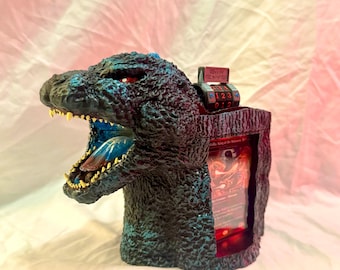 May include: A Godzilla-themed card holder, featuring a detailed sculpture of the iconic monster's head with an open mouth. The holder is dark blue with red accents, and includes a combination lock and a display area for cards.