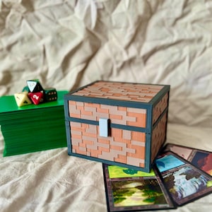 Puede incluir: Una caja de color marrón y negro con un patrón de ladrillo y un cierre gris. La caja está sobre una superficie beige con una pila de cartas de juego verdes y un montón de dados.