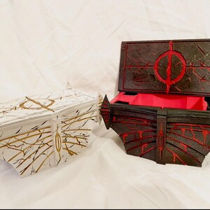 May include: Two decorative boxes. One is white with gold and black accents, the other is black with red accents. The black box is open, revealing a red interior. Both boxes have geometric designs.