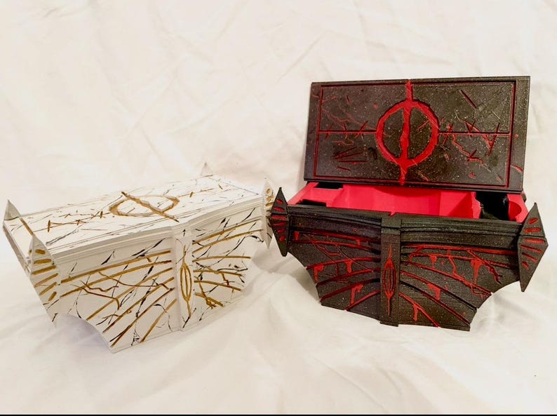 May include: Two decorative boxes. One is white with gold and black accents, the other is black with red accents. The black box is open, revealing a red interior. Both boxes have geometric designs.