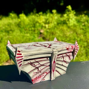 May include: A rectangular, white and gray chest with red accents. The chest has a geometric design with sharp, pointed edges and a textured surface. Red streaks and splatters add a dramatic effect to the chest's exterior.