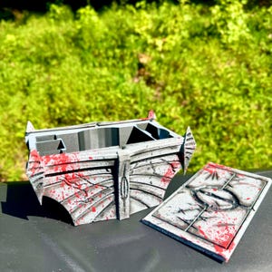 May include: A gray and white decorative box with a lid, featuring red splatter details. The box has an angular design with a central emblem and is set against a green, leafy background. The lid is separate and has a matching design.
