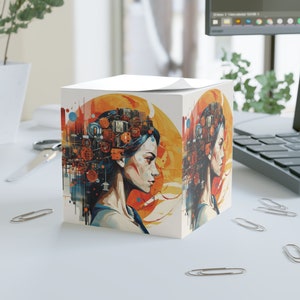 May include: A white cube-shaped notepad featuring a colourful illustration of a woman with a cityscape on her head. The cityscape is made up of buildings, gears, and other industrial elements. The woman's hair is blue and her face is partially obscured by the cityscape. The background is a gradient of orange, yellow, and white.