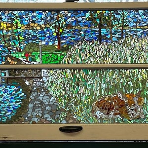 May include: A stained glass window depicting a lake scene with trees, water, and a grassy area. The window is divided into two sections by a horizontal bar. The top section shows a lake with trees on either side. The bottom section shows a grassy area with a small animal lying down.