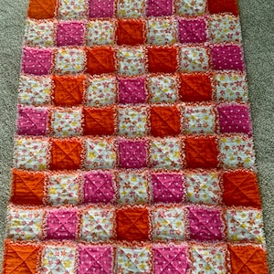 以下が含まれることがあります： オレンジ、ピンク、白の生地の正方形でできたパッチワークキルト。生地は花柄で、一部の正方形には白い水玉模様があります。キルトは縁がほつれています。