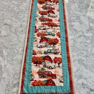 Op de afbeelding: Een rechthoekige, gewatteerde tafelloper met een herhalend patroon van rode schuren, vintage vrachtwagens en herfstbladeren. Het centrale ontwerp is omzoomd door een lichtblauwe stof en een rood geruite rand.