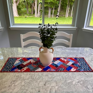 May include: A rectangular quilted table runner featuring a patchwork design in red, white, and blue fabric patterns. The runner has a red border and is centred with a matte cream-coloured ceramic vase containing green eucalyptus stems.