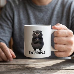 Könnte beinhalten: Ein weißer Keramikbecher mit einer Grafik eines mürrischen schwarzen Katers, der eine kleine blaue Fliege trägt und die Arme verschränkt hat. Unter dem Kater ist der Text "EW, PEOPLE" in einer fetten, schwarzen, serifenlosen Schrift gedruckt.
