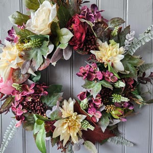 May include: A circular floral wreath with cream, burgundy, pink, and purple flowers, and green leaves. The wreath is densely packed with blooms and foliage, creating a full and textured appearance. Suitable for home decoration, it is a beautiful display.