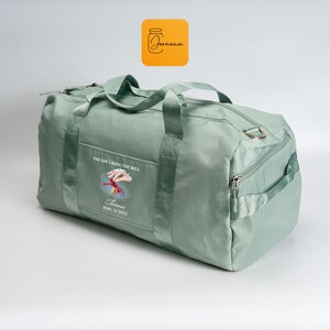 May include: A sage green duffel bag with handles and a shoulder strap. The bag has a front pocket with an embroidered design of a hand ringing a bell, with the text "THE DAY I RANG THE BELL" and "Thomas, May 21, 2021".