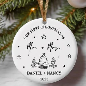 Peut inclure: Ornement de Noël en céramique blanche avec du texte noir qui dit "Our First Christmas as Mr. & Mrs. Daniel + Nancy 2023". L'ornement présente une illustration stylisée de deux sapins de Noël.