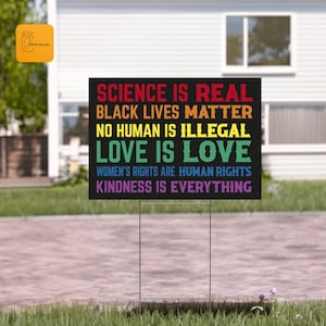 May include: A black yard sign with white text in a rainbow gradient. The text reads "Science is real, Black Lives Matter, No Human is Illegal, Love is Love, Women's Rights are Human Rights, Kindness is Everything."