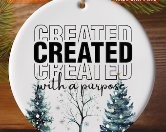 May include: White round Christmas bauble with a watercolour design of trees and snowflakes. The bauble has black text that reads "CREATED CREATED CREATED with a purpose".