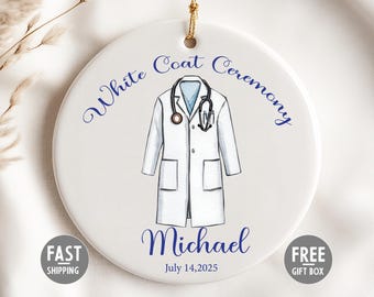 Könnte beinhalten: Weißes Keramik-Scheibenornament mit goldener Aufhängeschnur. Es zeigt eine Illustration eines weißen Mantels und eines Stethoskops. Der Text lautet "White Coat Ceremony" und "Michael July 14, 2025".