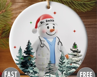 Könnte beinhalten: Weißer Keramik-Weihnachtsschmuck mit einem Schneemann, der als Arzt verkleidet ist und einen Weihnachtsmannhut mit dem Namen "Dr. Smith" trägt. Der Schneemann hat ein Stethoskop und ist von stilisierten Tannenbäumen und Sternen umgeben.