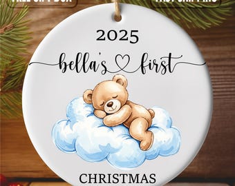Puede incluir: Adorno navideño de cerámica blanca con un oso de peluche durmiendo sobre una nube azul. El adorno tiene el texto "2025 bella's first CHRISTMAS". Se adjunta una cuerda para colgarlo.