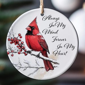 May include: A round, white ceramic ornament with a red cardinal bird illustration perched on a branch with red berries. The ornament has the text "Always In My Mind Forever In My Heart" in elegant script, and a jute string for hanging.