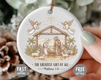 May include: A white ceramic Christmas ornament with a religious scene. The image depicts the nativity scene with angels, Mary, Joseph, and baby Jesus. Text reads "THE GREATEST GIFT OF ALL - Matthew 1:21".