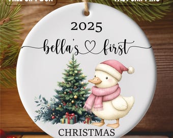 Puede incluir: Adorno navideño redondo de cerámica blanca con un pato con bufanda y gorro rosas, un árbol de Navidad y el texto "2025 bella's first CHRISTMAS". Incluye una ilustración de caja de regalo.