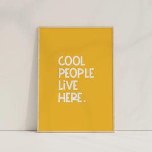 Op de afbeelding: Een gele ingelijste print met witte tekst die "COOL PEOPLE LIVE HERE." luidt.