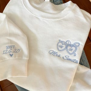 May include: White sweatshirt with light blue embroidered hearts and the text "Mrs. Smith". The left sleeve has "EST. 12.6.25" embroidered in light blue. The sweatshirt is folded on top of blue jeans.