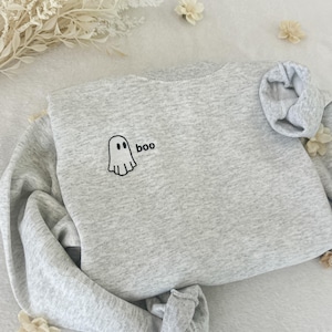 May include: A light gray sweatshirt with the word "boo" and a ghost graphic embroidered in black. The sweatshirt is folded, with the sleeves and bottom hem tucked in. Decorative white flowers and foliage are scattered around the sweatshirt.