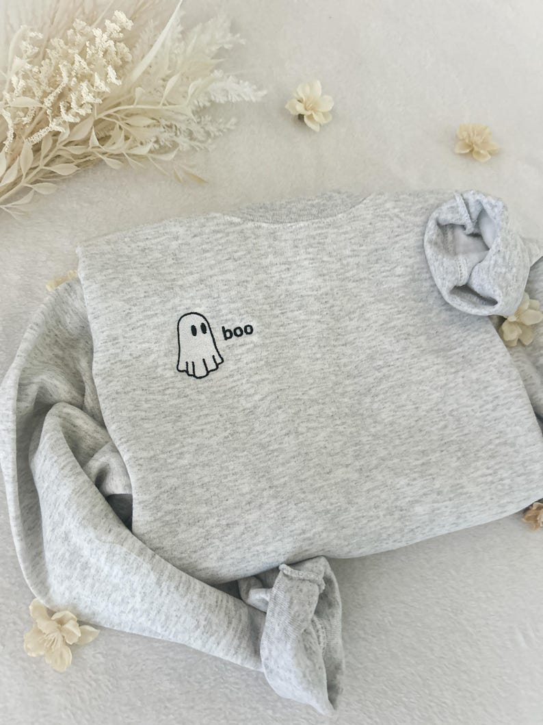 May include: A light gray sweatshirt with the word "boo" and a ghost graphic embroidered in black. The sweatshirt is folded, with the sleeves and bottom hem tucked in. Decorative white flowers and foliage are scattered around the sweatshirt.