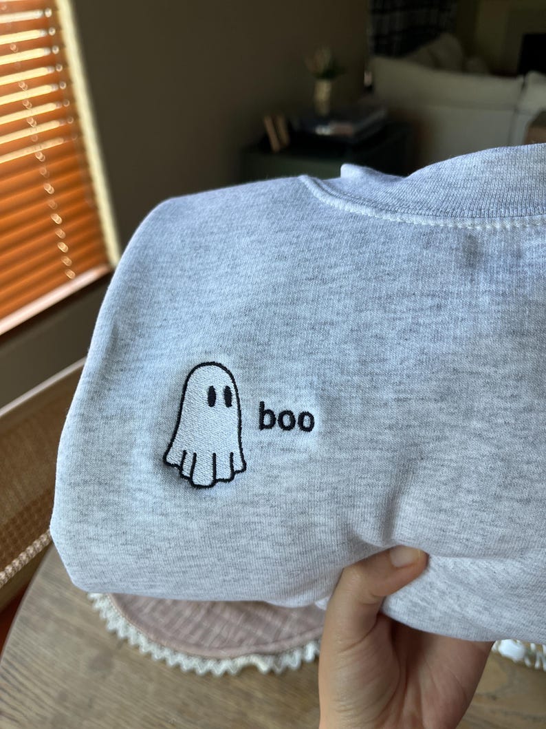 May include: A light gray sweatshirt with a small embroidered ghost and the word "boo" in black. The ghost has a simple, friendly design. The sweatshirt is folded, showing the design on the front.