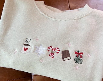 Pode incluir: Camisola creme com desenhos bordados com tema de feriado. Apresenta uma chávena de café, um floco de neve, bengalas de doces, um livro e azevinho. Acentos de estrelas cor-de-rosa estão espalhados pelos desenhos. A camisola tem gola redonda.