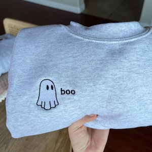 May include: A light gray sweatshirt with a crew neck. The sweatshirt features an embroidered ghost design with the word "boo" next to it. The ghost is white with a black outline and two black eyes. The text "boo" is in black.