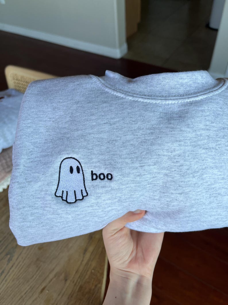 May include: A light gray sweatshirt with a crew neck. The sweatshirt features an embroidered ghost design with the word "boo" next to it. The ghost is white with a black outline and two black eyes. The text "boo" is in black.