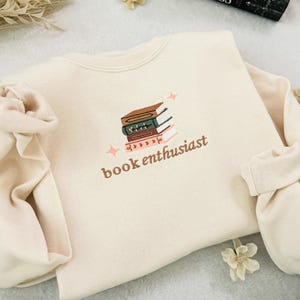 May include: Cream-colored sweatshirt with the embroidered text "book enthusiast" below a stack of books. The books are brown, green, and red. Two small pink stars are embroidered on either side of the books. The sleeves are gathered at the wrists.