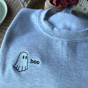 May include: A light gray sweatshirt with a small embroidered ghost and the word "boo" in black. The ghost has two black eyes and a smiling mouth. The sweatshirt is made of a soft material.