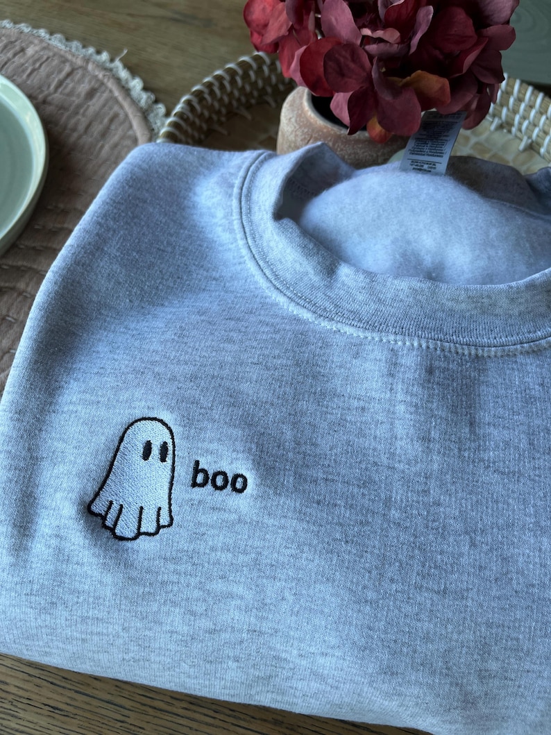 May include: A light gray sweatshirt with a small embroidered ghost and the word "boo" in black. The ghost has two black eyes and a smiling mouth. The sweatshirt is made of a soft material.