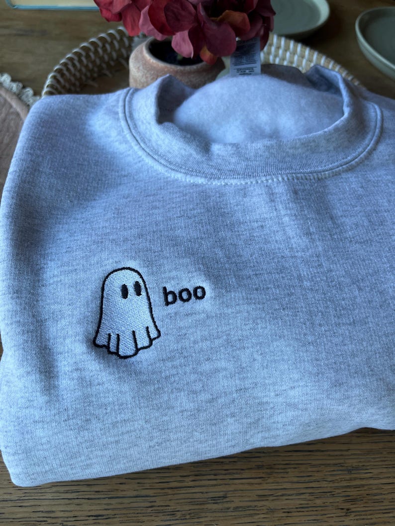 May include: A light gray sweatshirt with a crew neck. The sweatshirt features an embroidered ghost and the word "boo" in black. The ghost has two black eyes and a wavy bottom. The sweatshirt is made of a soft material.